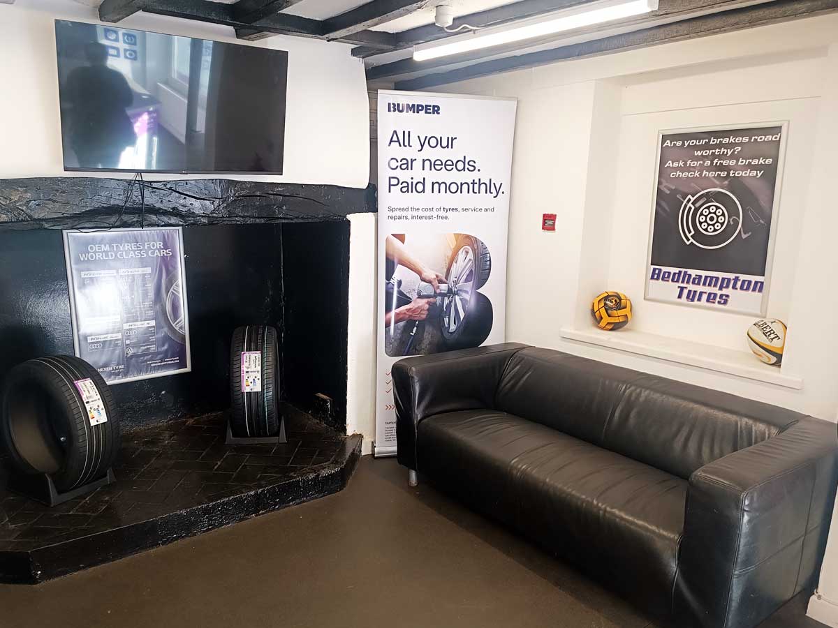 Internal view of the sitting area for waiting customers at Bedhampton Tyres showing a black sofa, tv on the wall and fireplace with various forms of advertising and posters on the wall